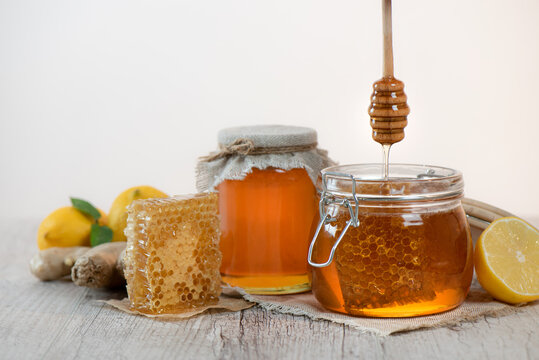 Honey in a jars, piece of bee comb and honey dipper on light background. Next to it is sliced lemons and ginger. Home treatment for flu. Honey harvesting concept.