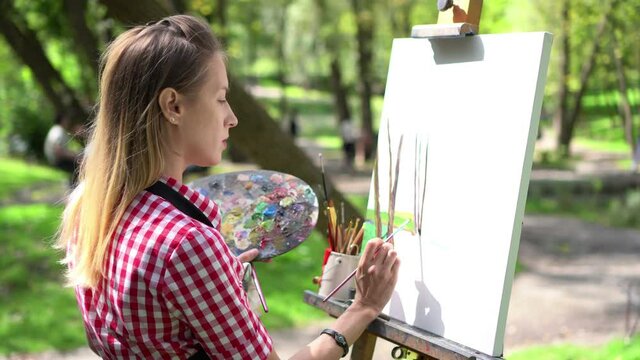 Young woman painting a picture with acrylic paints in the park. Canvas mounted on an easel. 
