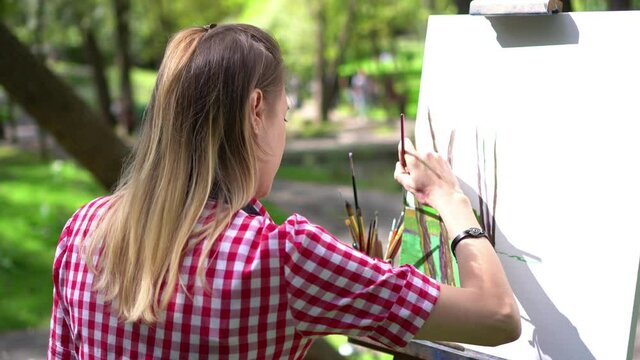 Woman artist at the plein air painting. She paints an oil painting on the canvas in the park. Her canvas stands on an easel. Girl holds a brush and palette in her hands. Creative mood.