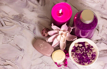 Composition of lilac sea salt bottle, soap bars, pink burning candle, lotus flower on white brushstrokes background flatly. Spa treatments, relaxation, recuperation, feminine concept. Copy space.