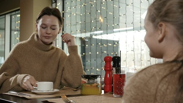 mother and daughter sit together in a restaurant and drink tea, talk against the background of a glowing garland. High quality FullHD footage