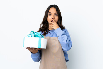 Pastry chef holding a big cake over isolated white background surprised and shocked while looking right