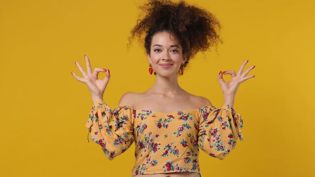 Spiritual tranquil smiling vivid young latin curly woman 20s wears casual flower dress hold spreading hands in yoga om aum gesture relax meditate try to calm down isolated on plain yellow background