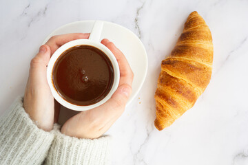 Top view of woman hands holding cup with hot chocolate drink and french croissant on marble table