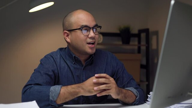 Young asian businessman working with computer video conference in the dark office at night. Attractive Indian man work hard overtime in home with floor lamp ambient warm light late at night
