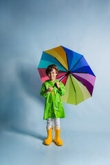 a little boy in a green raincoat and yellow rubber boots stands and holds a multi-colored umbrella on a blue background with a place for text