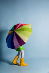 a little boy in a green raincoat and yellow rubber boots covered himself with a multicolored umbrella on a blue background with a place for text
