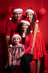 Fototapeta premium Hermosa familia de cuatro integrantes festejando la navidad en un fondo rojo con regalos y gorros navideños viendo a cámara