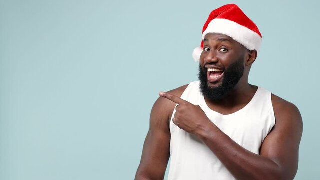 Merry promoter young african Santa man in white tank top Christmas hat pointing finger aside workspace copy space mockup promo commercial area isolated on plain light blue background studio portrait