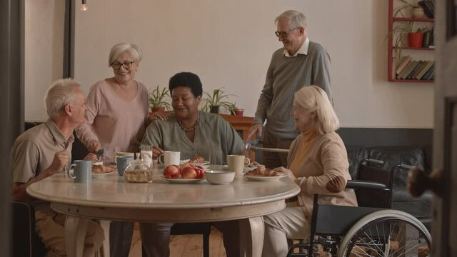 Slowmo medium portrait of group of happy senior men and women waving and smiling to camera sitting around dining table in nursing home