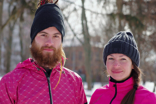 Cheerful Couple Wearing Black And Pink Sportswear Looking At Camera, Winter Park Behind Them. Young Man And Woman Posing Outside Together. Concept Of Healthy Lifestyle