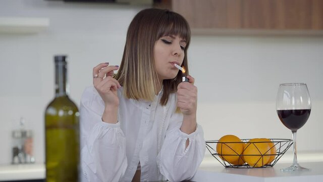 Medium shot portrait of slim beautiful young woman lighting cigarette smoking in kitchen at home. Sad Caucasian millennial person with wine bottle indoors thinking. Slow motion