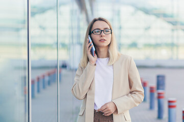 Fototapeta premium portrait of a serious woman wearing glasses blonde holding a mobile phone talking on a smartphone near the office