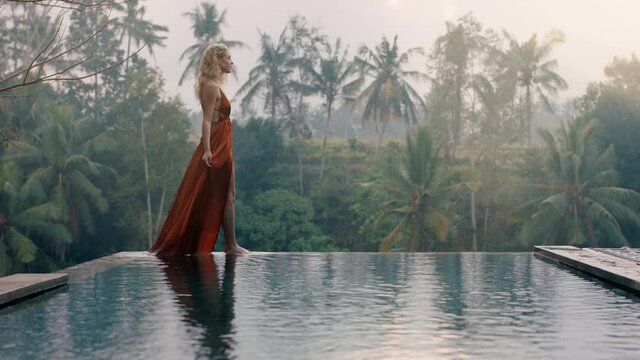beautiful woman walking by infinity pool wearing elegant dress enjoying luxury lifestyle on exotic vacation at hotel spa with view of tropical jungle in morning mist