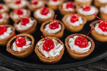A lot of dough tartlets close-up with cream, cherries stand on a round tray, a plate on a wooden table. Banquet, buffet, photography