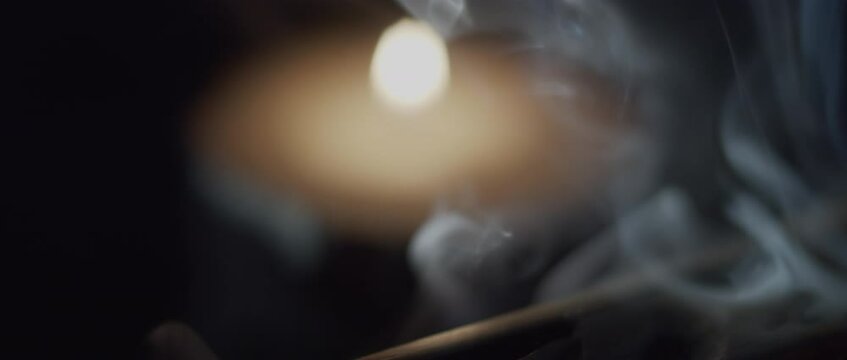 Close up of incense stick burning in a wooden holder, candle in the background. Slow motion, shallow depth of field. 