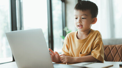 handsome cute Asian boy with looks at the camera while sitting and studying at home on a laptop to...