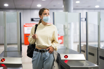 Woman traveler with long hair in protective face mask passing the turnstiles at subway station