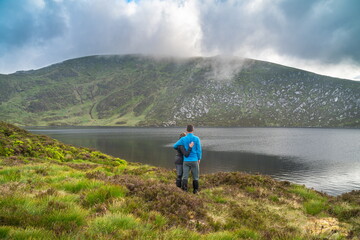 Happy backpackers. Young hipster man woman couple standing, lakeshore, beside a lake with epic, scenic mountain background. Wicklow, Ireland