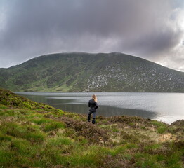 Fototapeta na wymiar Happy backpackers. Young hipster woman standing, lakeshore, beside a lake with epic, scenic mountain background. Wicklow, Ireland
