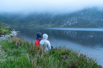 Happy backpackers. Young hipster man woman couple sitting lakeshore, beside a lake with epic, scenic mountain background