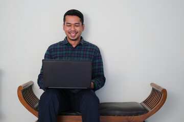 Young Asian man sitting in the wooden couch while working with his laptop showing happy face expression