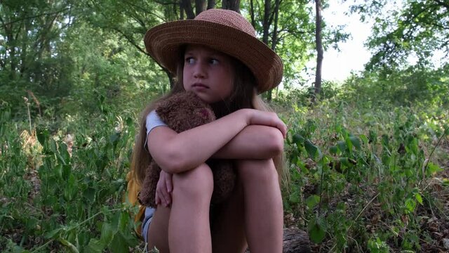 Scared girl with a toy bear and yellow backpack sits alone in the thicket. Child tourist lost in the forest. Upset kid waiting for her to be found in woods. Close up view