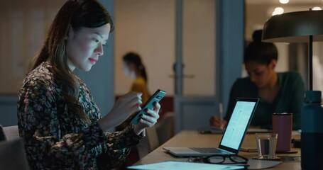 asian business woman working late using laptop computer in office sitting at desk checking smartphone brainstorming ideas on mobile technology