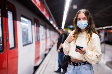 Portrait of a thoughtful girl in a protective mask, standing on the platform of a subway station...