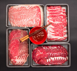 Sliced raw beef place in a row on a black plastic tray for cooking, fresh meat for grilling, yakiniku, sukiyaki or shabu. Chopsticks are placed on a bowl of sauce.