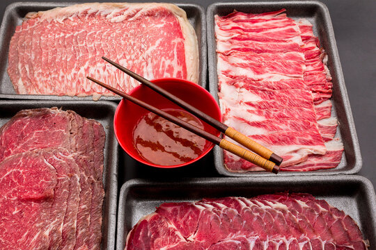Sliced raw beef place in a row on a black plastic tray for cooking, fresh meat for grilling, yakiniku, sukiyaki or shabu. Chopsticks are placed on a bowl of sauce.