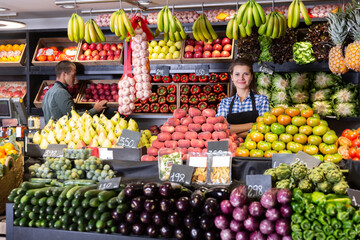 .Smiling woman seller stands near counter with fresh fruits and vegetables at a grocery store.