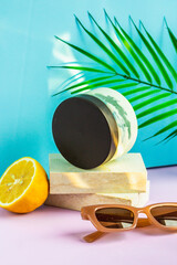 Cosmetics are on the podium on a summer background, in the rays of the sun. a jar of cream is on the podium, half a lemon and sunglasses are lying next to it, and a palm leaf is hanging above it