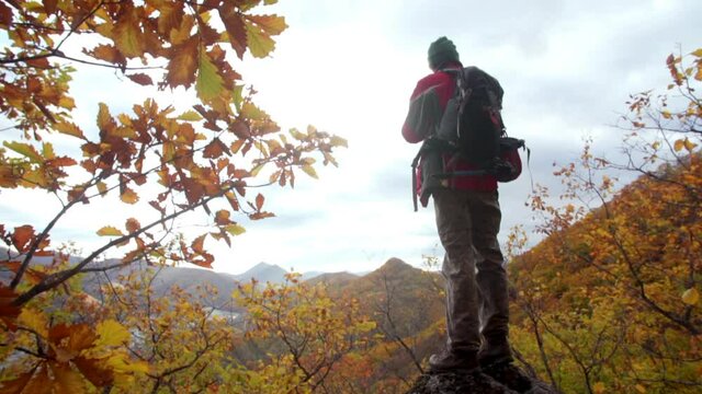 Back of a photographer standing on a high rock and taking pictures of a small city situated below in a valley between mountains. Autumn