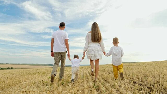 Farmer family with children sons walks joining hands among field on mown wheat on sunny day against blue sky and forest on horizon backside view