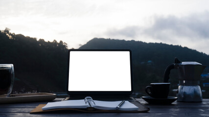 Fototapeta premium Computer tablet, notebook, moka pot and coffee cup on wooden table with mountain view in background.