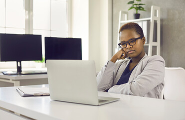Serious woman sitting at office desk and looking at screen of modern laptop computer with thoughtful face expression. Concentrated black business lady thinking over complicated task, idea, new project
