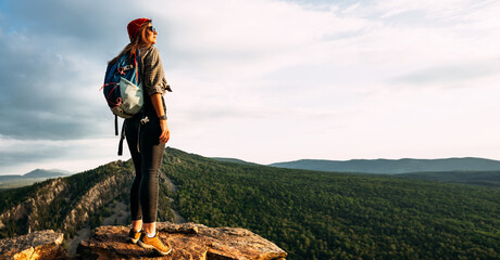 A young female tourist with a backpack admires the sunset from the top of the mountain, panorama. A traveler on the background of mountains. Tourist with a backpack on the background of mountains