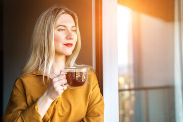 Beautiful smiling woman standing near the window and drinking tea cup. Female pretty face, work from home, social distancing, alone at home in office. close up front portrait
