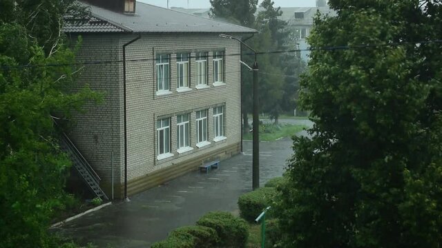 Bad weather. It is raining heavily. Water is pouring from drainpipes on the building. The trees are swaying.