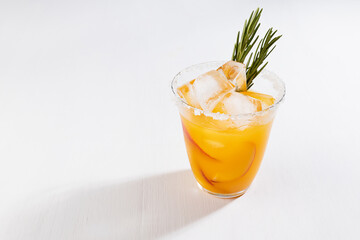 Fresh cold peach drink in glass with ice cubes, green rosemary, fruit slice, sugar rim on soft light white wooden table, top view.