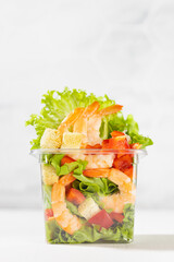 Organic vitamin meal - fresh sea food salad with shrimps, lettuce, red bell pepper, croutons in plastic box in white restaurant interior with marble tile wall, vertical.