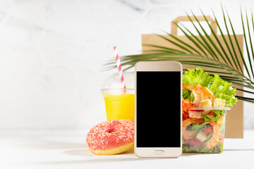 Fitness food set of orange juice, shrimp salad in box, donut, packet, blank phone in white interior with green palm leaf. Mockup advertising for delivery service, take away restaurant, online order.