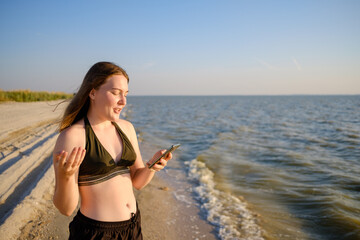 A young woman on the sea beach communicates via video link using a smartphone. Modern communication technologies. The girl is lying a swimsuit. Sunny summer day.