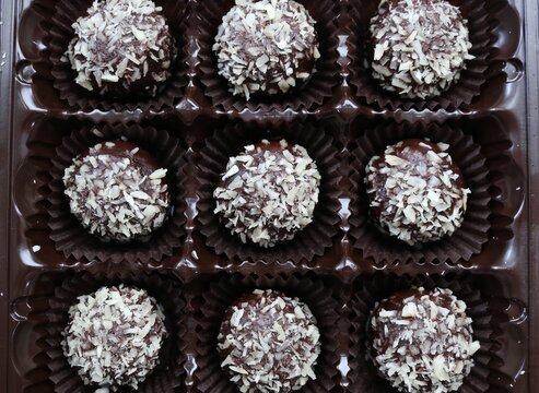 sweet candy contrasting background full frame, top-angle view of an open box of chocolate balls in coconut flakes lying in individual cells in plastic packaging, truffle mini cakes in hazelnuts