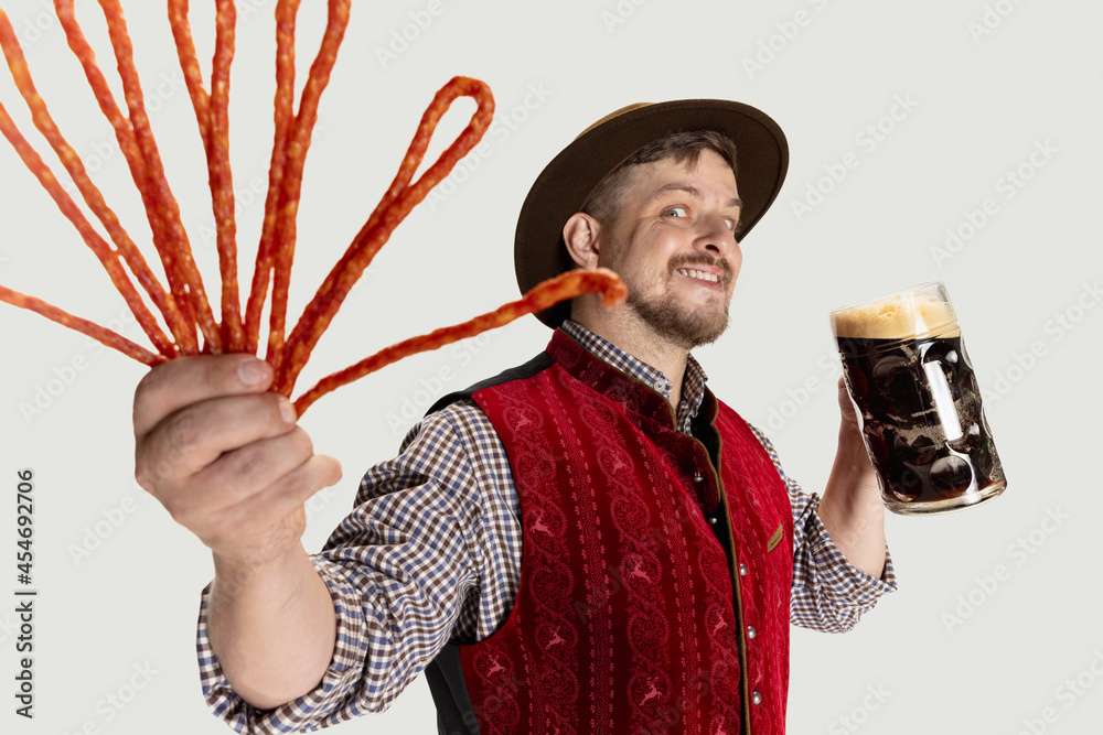 Wall mural Close-up of bearded man, waiter in traditional Austrian or Bavarian costume holding mug, glass of dark black beer isolated over gray background