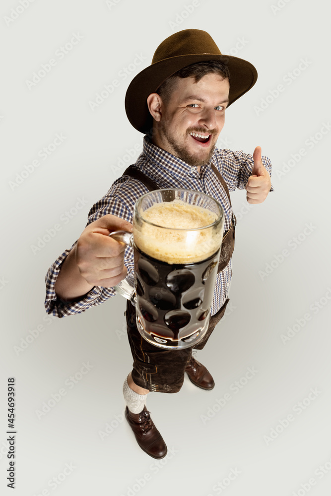 Wall mural High angle view of bearded man, waiter in traditional Austrian or Bavarian costume holding mug, glass of dark black beer isolated over gray background