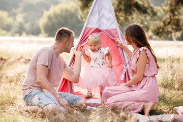 Naklejka na ściany i meble Mom, dad and little daughter are sitting next to wigwam decoration in the park. Family spending time outdoor in summer, having fun together. Girl are dressed in pink dress