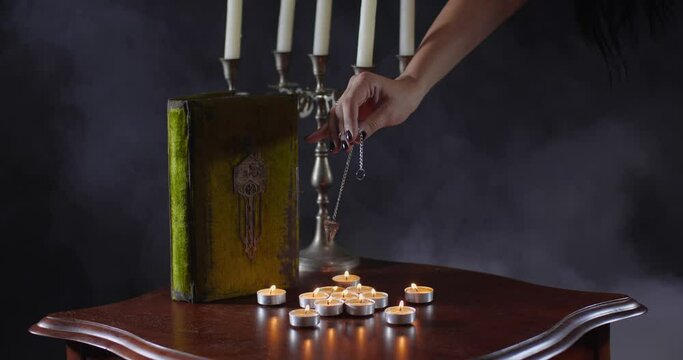 Close-up detailed shot of fortune-teller's hands with pendulum hovering over table with candles and old book on black background with smoke. Book of dead.  Predicting future or Halloween concept
