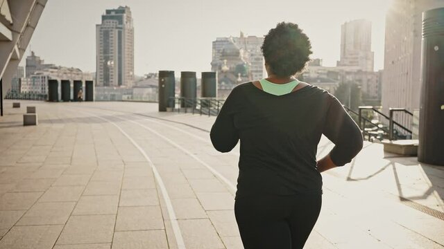 Morning jog and weight loss concept. Back view of overweight african american woman running in city area, sun flare